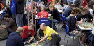 Erwachsene und Jugendliche sitzen an Tischen und auf dem Boden und spielen Brettspiele an einem Messestand von Ravensburger