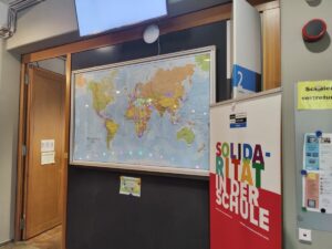 Weltkarte mit kleinen Leuchten hängt an Wand in Schulgebäude. Rechts davon ein Plakat.