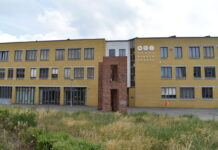 Ein gelbes zweistöckiges Schulgebäude mit einem Kunstwerk aus rotem Backstein davor steht hinter einem Grünstreifen: Rahel-Hirsch-Schule