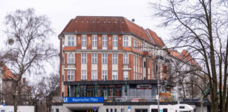 Rot-weißer Altbau steht hinter dem Eingang zum U-Bahnhof Bayerischer Platz in Berlin. Davor Wiese und zwei kahle Bäume.