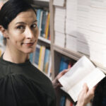 Frau mit dunklen Haaren und dunklem Kleid steht vor Bibliotheksregal, mit offenem Buch in den Händen, schaut nach rechts in Kamera