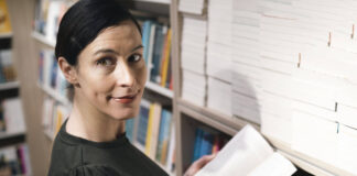 Frau mit dunklen Haaren und dunklem Kleid steht vor Bibliotheksregal, mit offenem Buch in den Händen, schaut nach rechts in Kamera