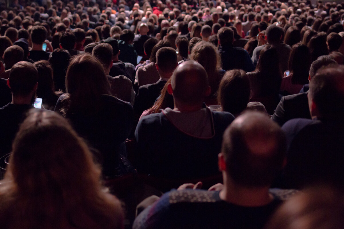 In the dark hall there is a view from the back of a crowd of hundreds of people sitting and watching the screen in a movie theater or performing on stage. Volle Säle: Darauf haben die Berliner Bühnen und Opernhäuser lange gewartet. Bild: iStock/Getty Images Plus/Ksenia Lyubasova