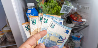 Eine Hand hält Euro-Scheine vor Lebensmitteln im vollen Kühlschrank. Foto: IMAGO / Bihlmayerfotografie