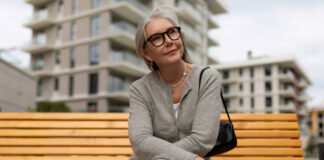 Lächelnde Frau mit grauem Haar und moderner Brille sitzt auf einer Holzbank. Bild: IMAGO / Zoonar